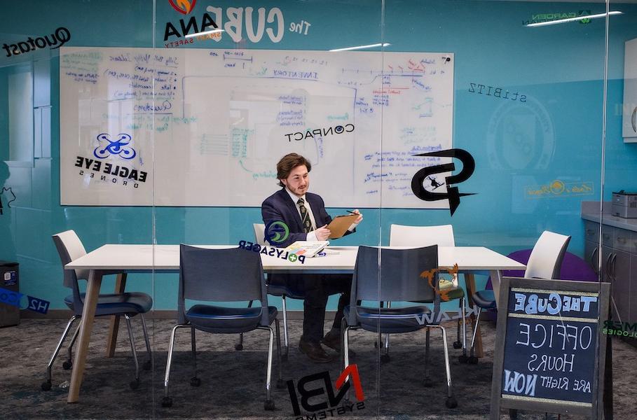 A man at Clarkson University sits inside a glass-walled room named the Cube while preparing business plan information using a clipboard.